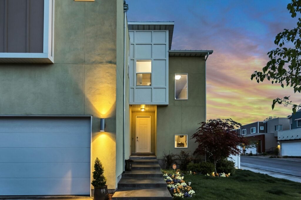 Modern house exterior with warm lighting and a landscaped garden at sunset.